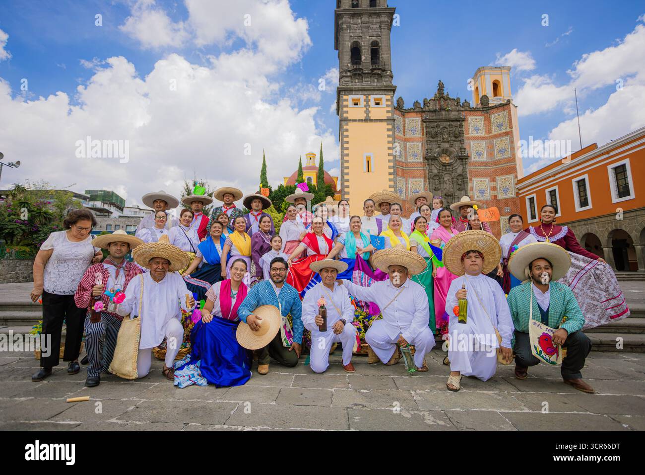 Eine große Gruppe von Tänzern und Menschen in traditionellen mexikanischen Kostümen posiert vor einer historischen Kirche unter einem blauen Himmel mit Wolken. Stockfoto