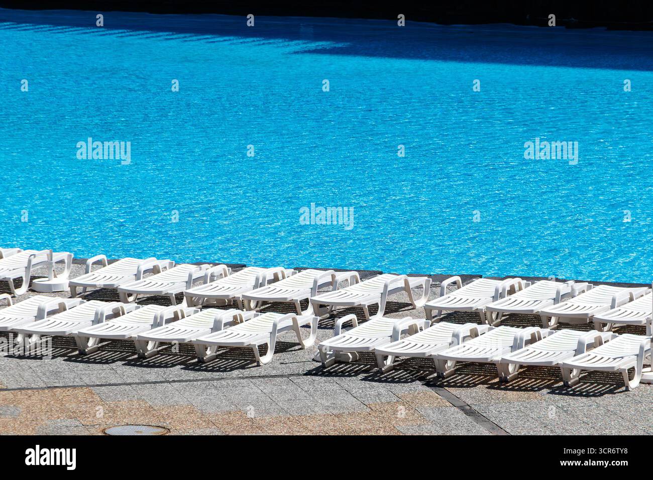 Leerer Pool mit zahlreichen weißen Sonnenliegen, die an einem sonnigen Tag neben einem großen, ruhigen blauen Swimmingpool stehen Stockfoto