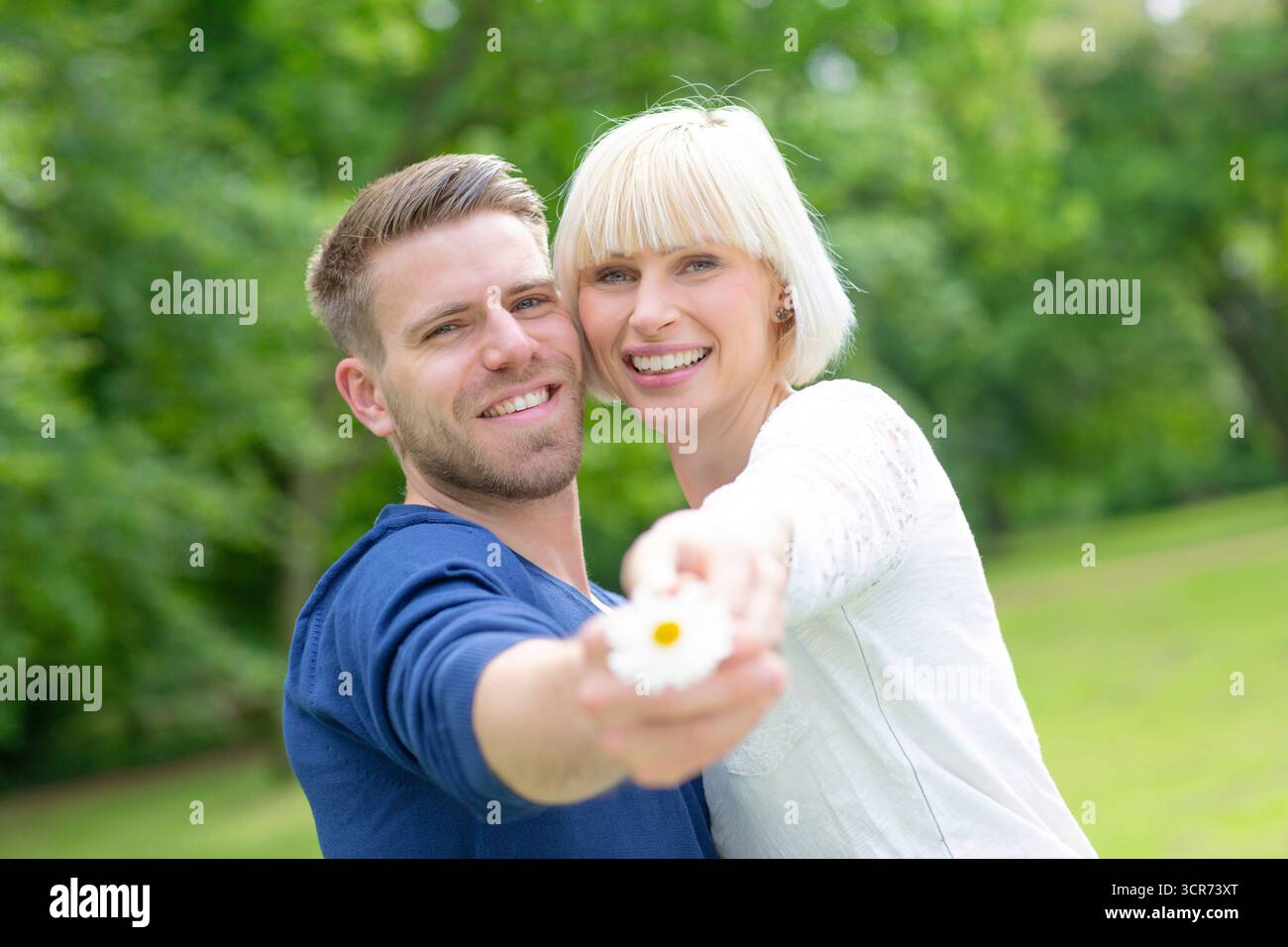 Ein verliebtes Paar steht glücklich auf einer Wiese und hält ein Gänseblümchen für die Kamera zusammen. Stockfoto