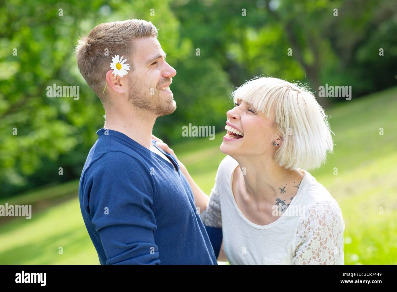 Ein verliebtes Paar steht auf einer Wiese. Sie hat ein Gänseblümchen hinter seinem Ohr versteckt und lacht Stockfoto