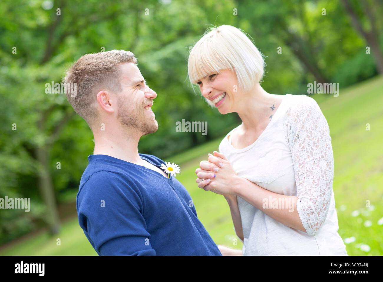 Ein verliebtes Paar steht auf einer Wiese. Sie steckt ihm ein Gänseblümchen in den Revers und macht so viel Spaß. Er grinst. Stockfoto