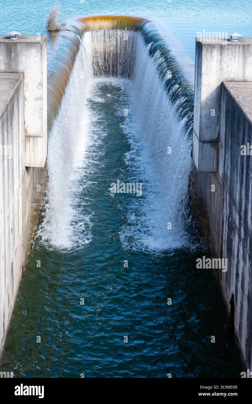 Wasser fließt über eine Betonbarriere und erzeugt einen Wasserfalleffekt in einer ruhigen Umgebung unter klarem Himmel. Stockfoto