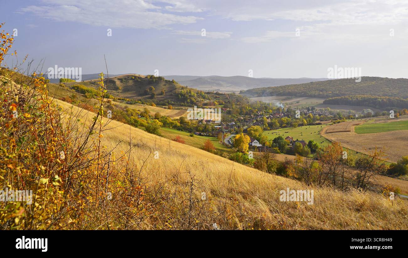 Ländliche Landschaft im Herbst. Blick auf das Dorf in den Bergen, Siebenbürgen. Sanfte Hügel, die mit goldenem Herbstgras bedeckt sind, führen zu einer malerischen Landschaft. Stockfoto