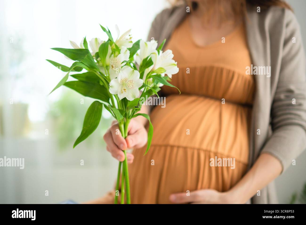Sie erwartet Mutter, die sanft weiße Blumen hält, während sie ihren Baby-Buckel berührt. Weiches natürliches Licht verstärkt diesen zarten mütterlichen Moment zu Hause. Stockfoto