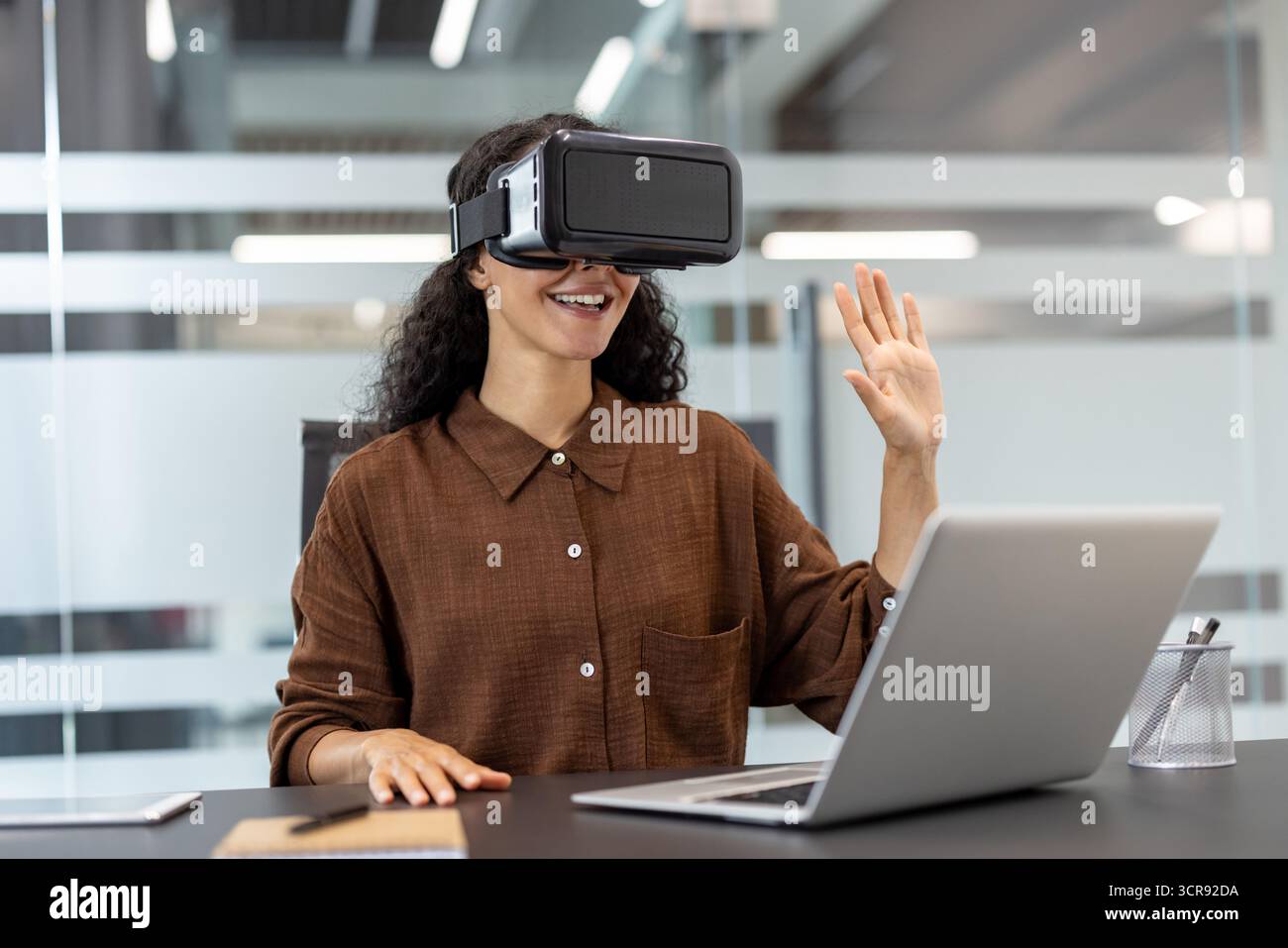 Junge Frau, die ein vr-Headset trägt, lächelt und Gesten macht, während sie in einem modernen Büro an einem Laptop arbeitet und in die virtuelle Realität und Metaverse-Technologie eintaucht Stockfoto