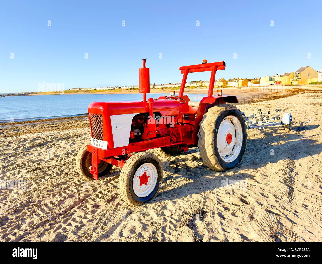 Roter Traktor, der an einem Sandstrand mit blauem Himmel und einem Küstendorf im Hintergrund in der Nähe des Ozeans geparkt wurde Stockfoto
