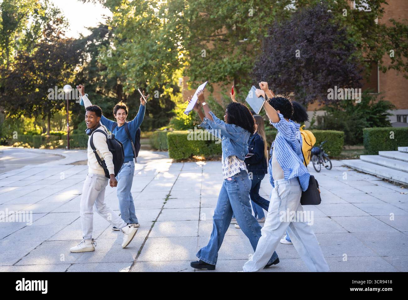 Verschiedene Gruppen junger Universitätsstudenten, die nach dem Unterricht auf dem Campus spazieren gehen und glücklich die Arme heben, um akademische Leistungen und Freundschaft zu feiern Stockfoto