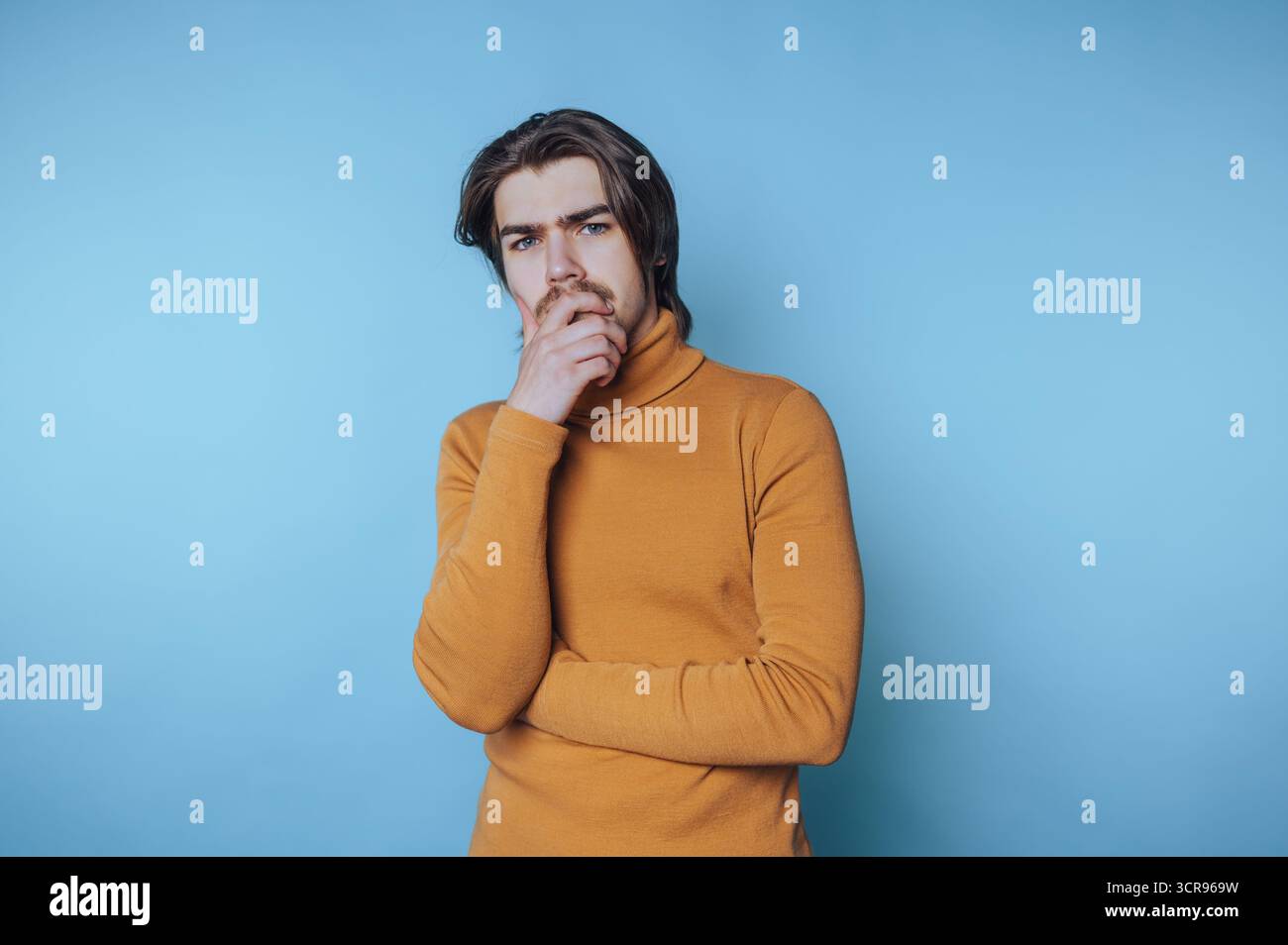 Der junge Mann steht mit einer nachdenklichen Pose, Hand am Kinn und reflektiert tiefgründige Ideen und Konzepte auf blauem Hintergrund Stockfoto