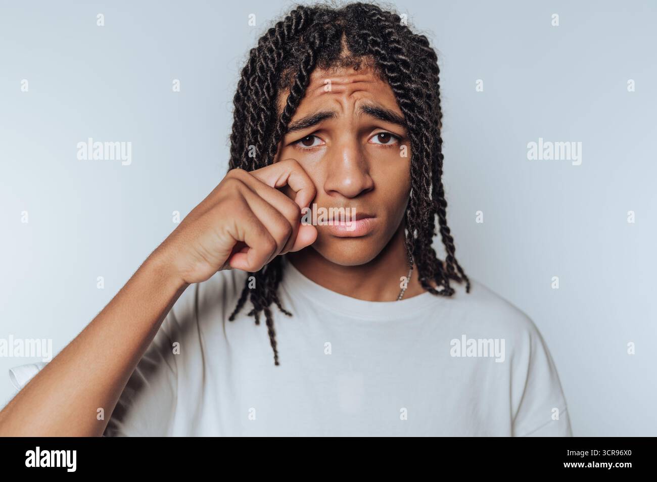 Ein trauriger junger Mann wischt sich sein Auge mit einer sanften Geste ab, die das Wesen persönlicher Reflexion und Verletzlichkeit auf weißem Hintergrund einfängt Stockfoto