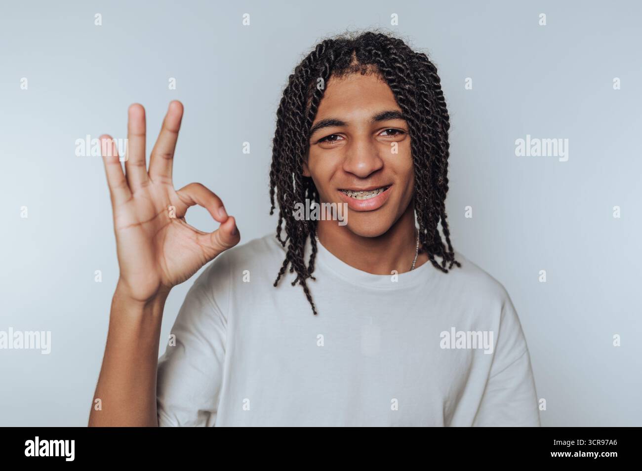 Der junge Mann blitzt mit einem Lächeln auf ein „OK“-Zeichen und zeigt Selbstvertrauen und Positivität auf weißem Hintergrund Stockfoto