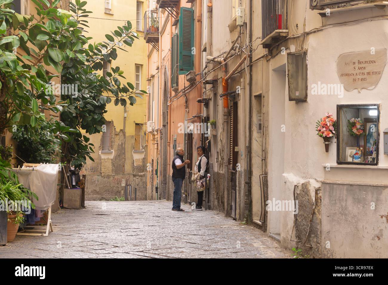 Mann und Frau stehen in einer engen Kopfsteinpflastergasse in Neapel mit einem traditionellen Straßenschrein (edicola votiva), Italien. Stockfoto
