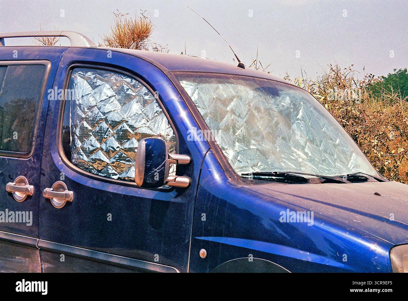 Silberner Sonnenschirm im Inneren eines blauen Autos, das neben Büschen geparkt ist, fotografiert als authentischer Analog-Schnappschuss mit starken Reflexionen und alltäglichem Sommercharakter Stockfoto