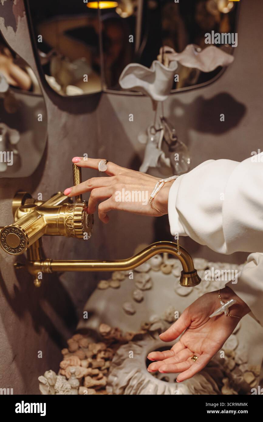 In einem stilvollen Raum wäscht sich eine junge Frau mit einzigartigen Perlen am Handgelenk ihre Hände in einem dekorativen Waschtisch. Das Becken ist mit Muscheln ausgestattet, Addin Stockfoto