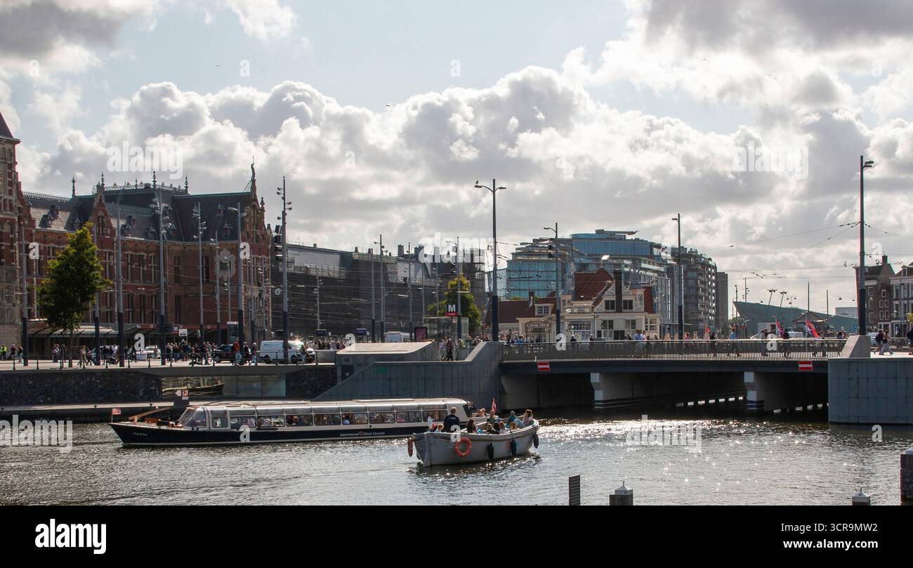 Amsterdam, Niederlande - 5. August 2025: Ein Boot gleitet durch ruhiges Wasser in der Nähe moderner Gebäude unter bewölktem Himmel. Stockfoto