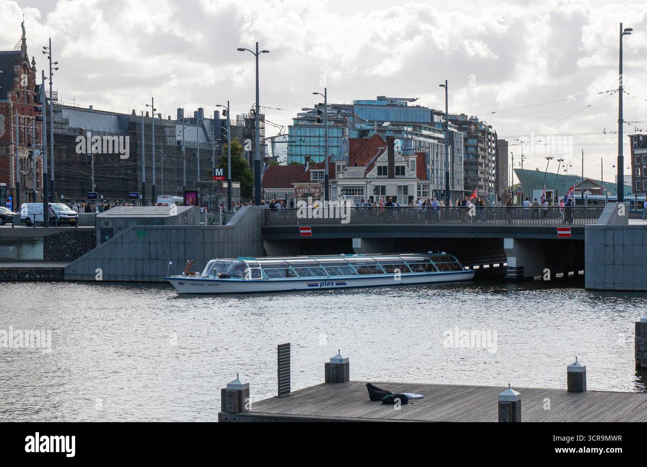 Amsterdam, Niederlande - 5. August 2025: Ein Boot gleitet unter einer Brücke, während die Menschen entlang der nahe gelegenen Uferpromenade spazieren. Stockfoto