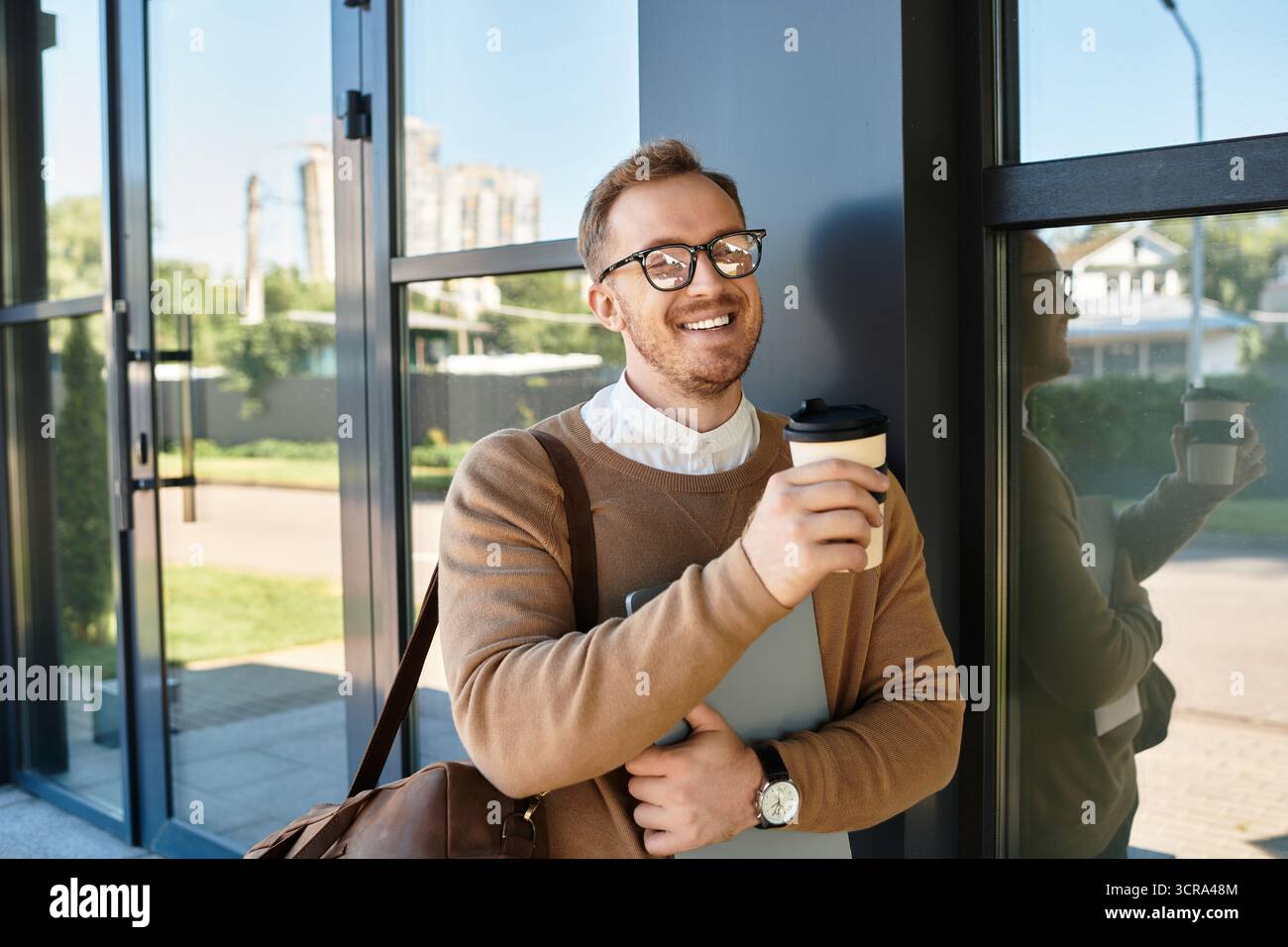 Ein junger Mann in einem Pullover steht vor einer modernen Universität, hält Kaffee und ein Notizbuch. Stockfoto