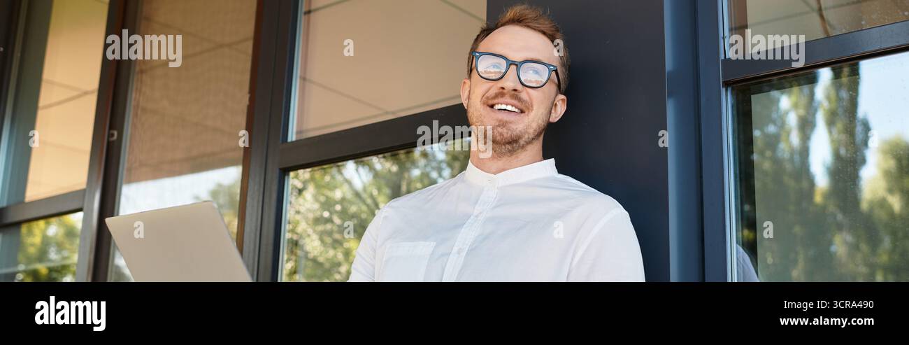 Ein junger Hochschullehrer genießt einen Moment der Freude, während er neben großen Fenstern steht. Stockfoto