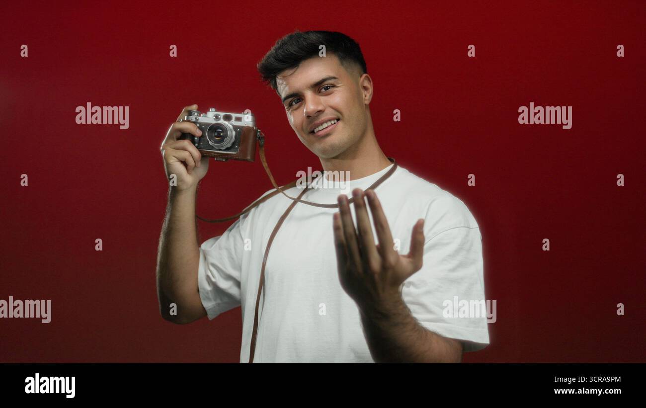Junger Mann mit einer alten Kamera, der eine Geste über isoliertem rotem Hintergrund macht und einen selbstbewussten und einladenden Ausdruck zeigt. Stockfoto