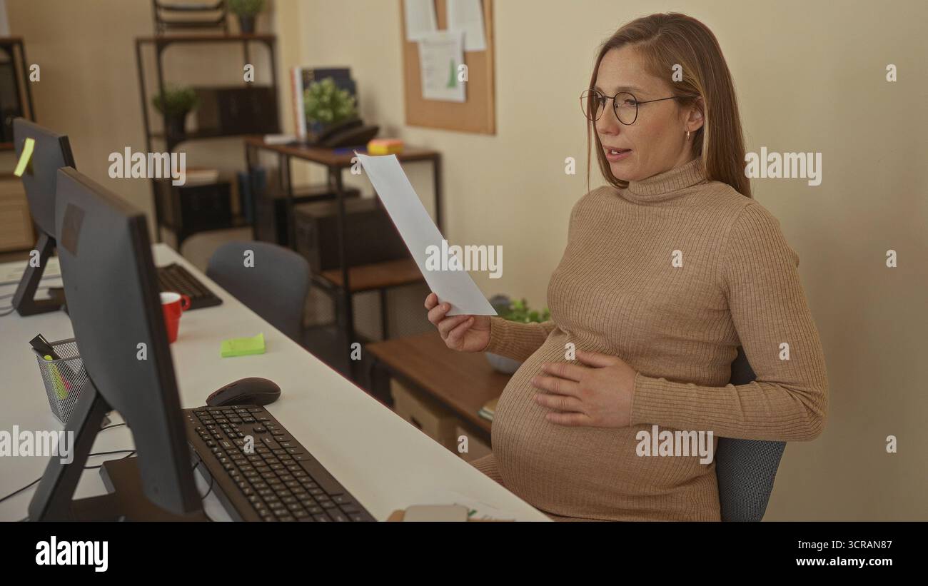 Schwangere Frau, die im Büro sitzt und Dokumente hält, am Schreibtisch arbeitet, von Computern umgeben ist und die Arbeitsumgebung betont. Stockfoto