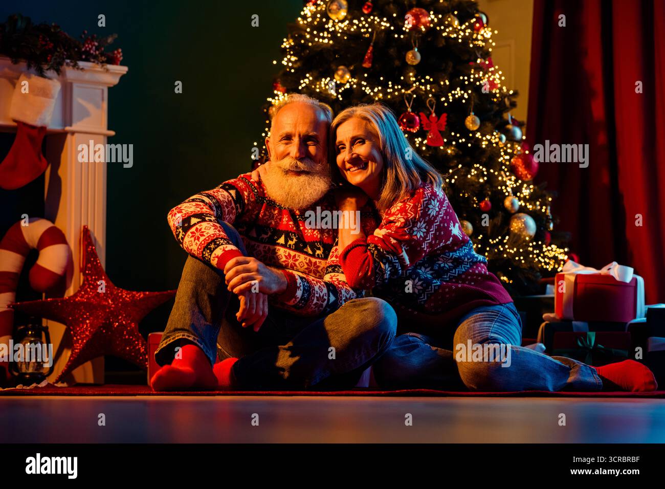Ein liebevolles älteres Paar sitzt in der Nähe eines wunderschön dekorierten Weihnachtsbaums in einer gemütlichen Atmosphäre mit Geschenken und warmen Lichtern, die eine festliche Stimmung schaffen Stockfoto