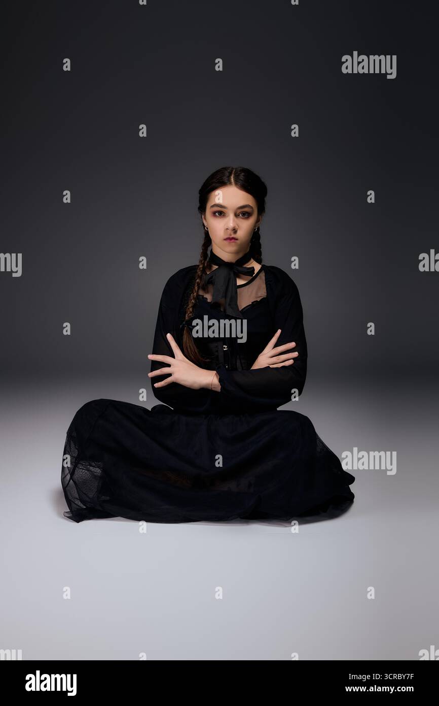Ein Mädchen mit Zöpfen sitzt im Kreuz und trägt ein gotisches schwarzes Mittwoch-Outfit und feiert Halloween. Stockfoto