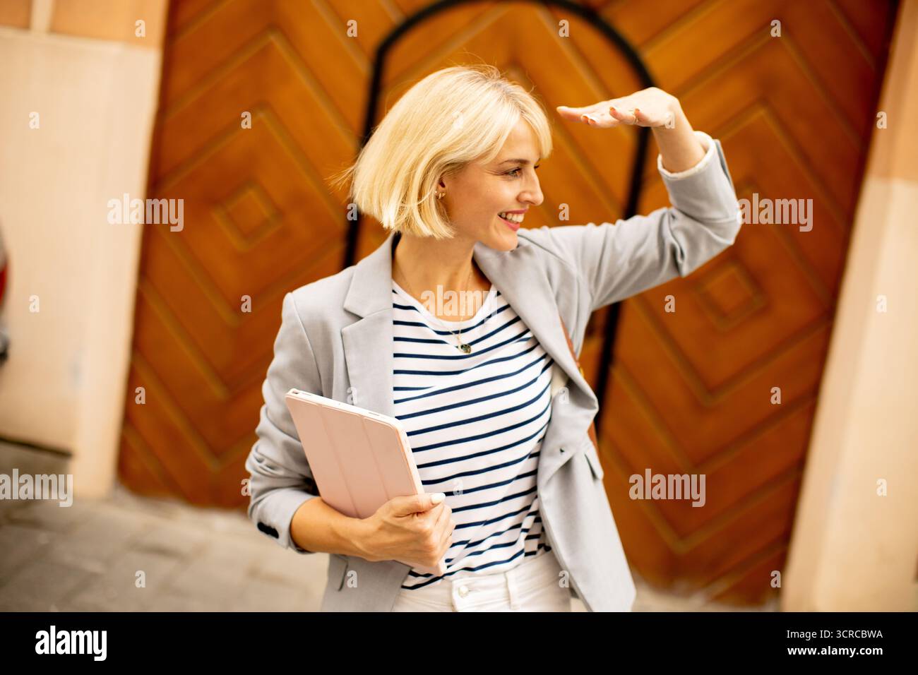 Eine Frau mit kurzen blonden Haaren lächelt, während sie ihre Augen vor hellem Sonnenlicht schützt. Sie hält ein Tablet in einer Hand und trägt ein lässiges Outfit Stockfoto