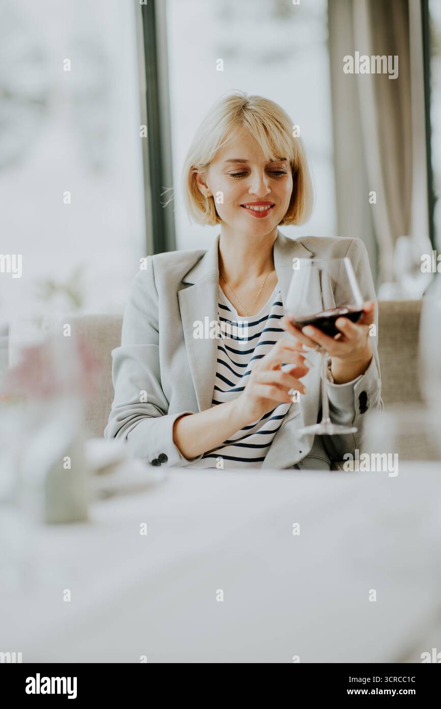 Eine Frau mit kurzen blonden Haaren lächelt, als sie ein Glas Rotwein untersucht. Sie sitzt an einem gut gedeckten Tisch in einem modernen Restaurant, umgeben von sanften Long Stockfoto