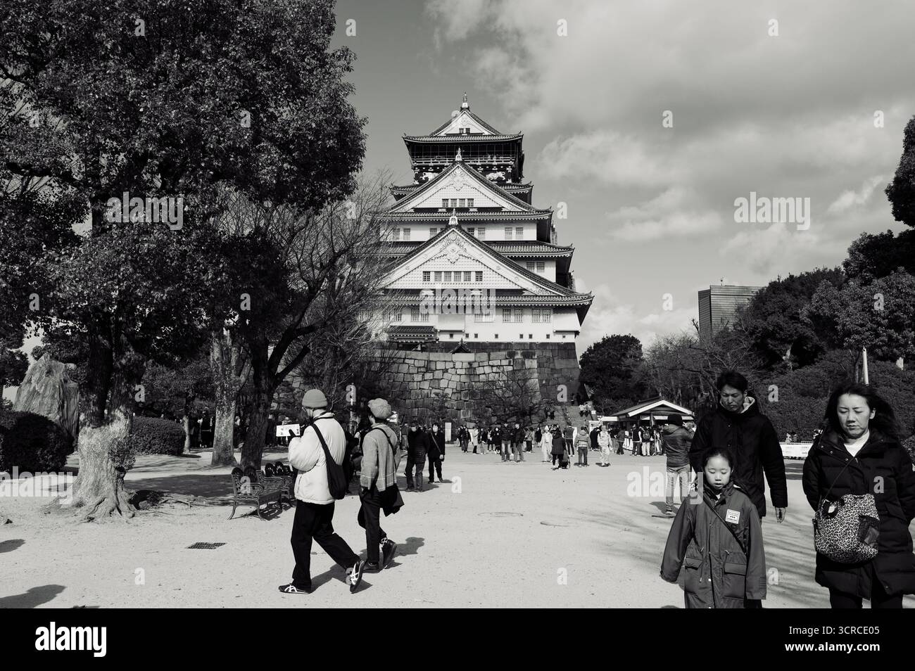 Besucher schlendern durch das Gelände des Schlosses Osaka, dessen großartige Architektur von Bäumen umrahmt wird und einen teilweise bewölkten Himmel Stockfoto