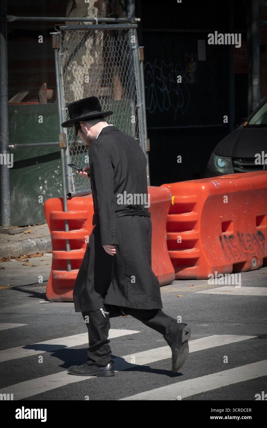 Ein nicht identifizierbarer jüdischer junger Mann überquert die Straße, während er auf sein Klapptelefon blickt. In Williamsburg, Brooklyn, New York. Stockfoto