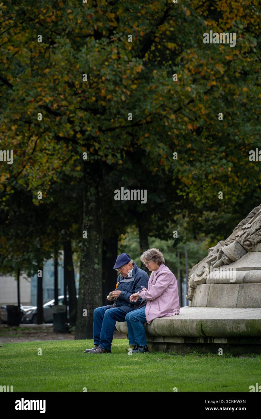 Älteres Ehepaar, das sich in einem Park in Porto, Portugal, auf dem Steinfuß des Denkmals für die Helden des Halbinsel-Krieges entspannt und sitzt Stockfoto