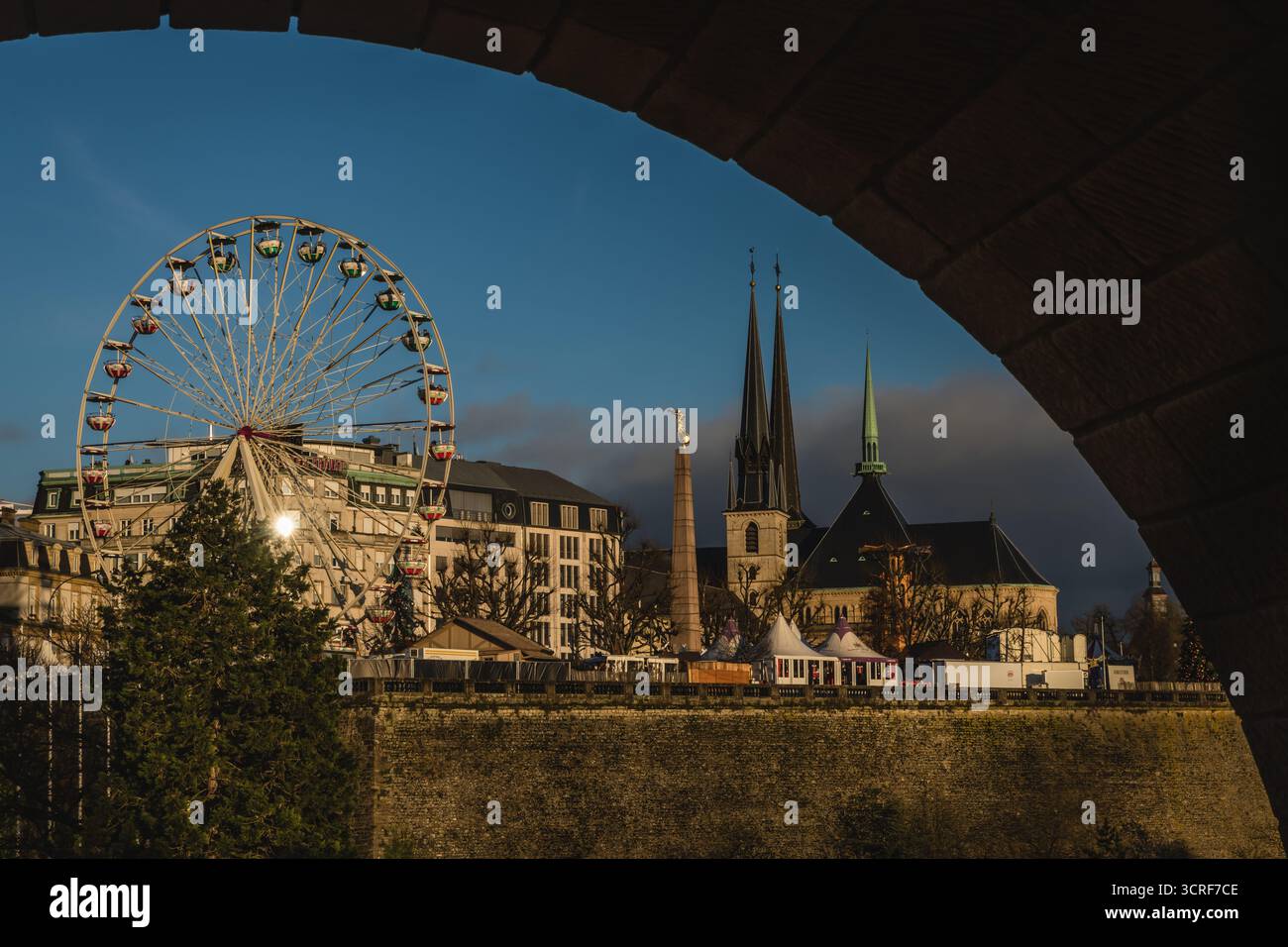Blick auf ein pulsierendes Riesenrad und die scharfen Kirchtürme ragen über die Skyline der Stadt unter einem Steinbogen, Luxemburg-Stadt. Stockfoto
