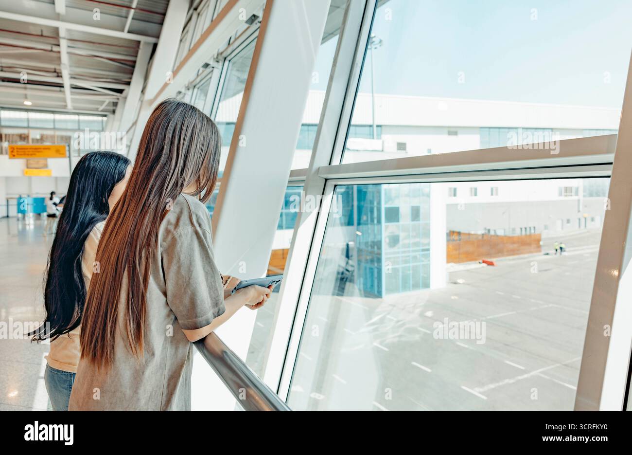 Zwei junge Frauen, eine mit langen schwarzen Haaren und die andere mit langen braunen Haaren, stehen an einem großen Fenster an einem Flughafen und blicken auf die Landebahn. Stockfoto