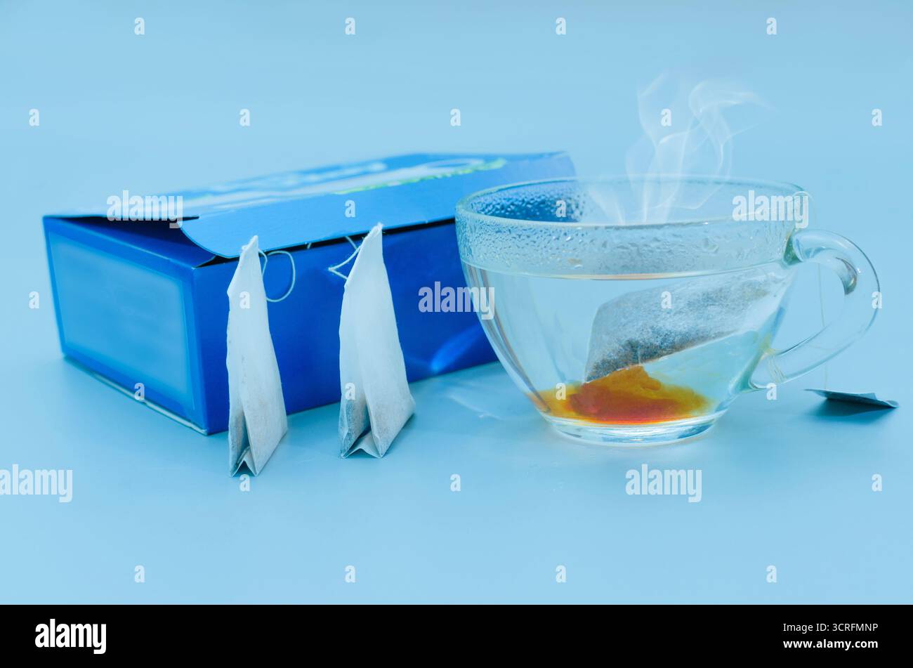 Eine einfache, einladende Stilllebensaufnahme mit einer klaren Glasschale heißen Tees, die die satte Farbe des Infusionsmittels zeigt. Zwei unbenutzte Pyramiden-Teebeutel Stockfoto