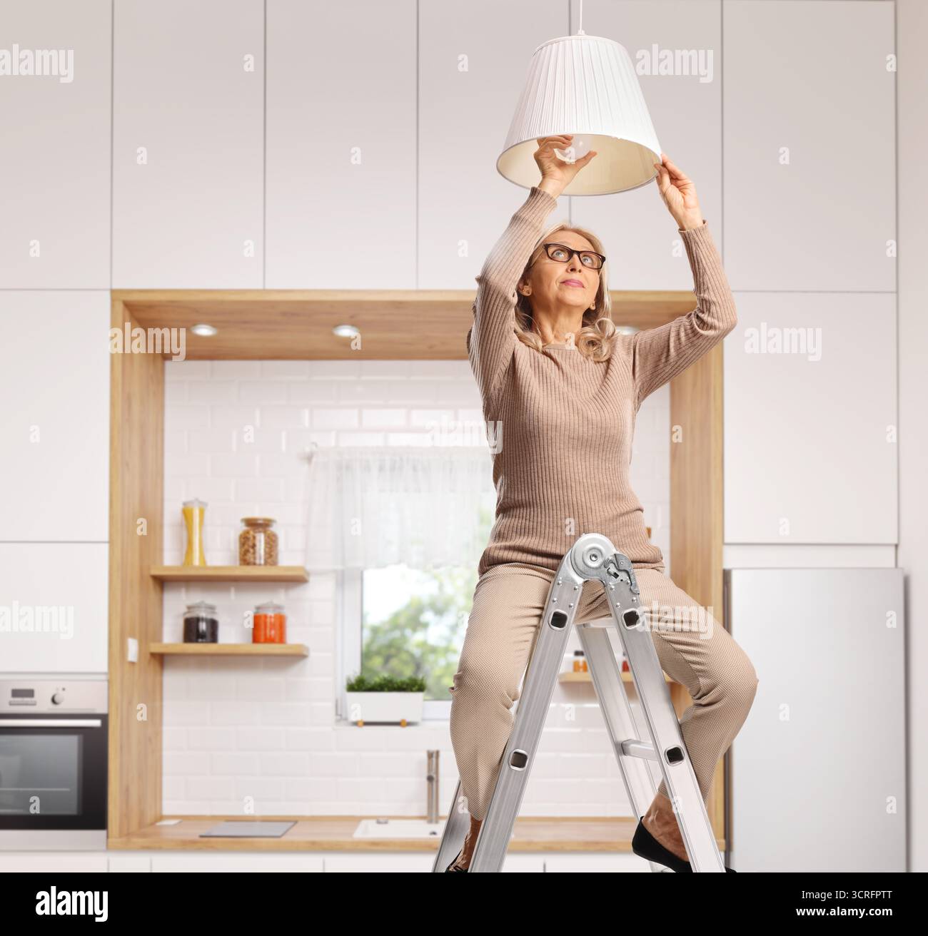 Reife Frau, die auf einer Leiter sitzt und eine Pendellampe in einer Küche wechselt Stockfoto