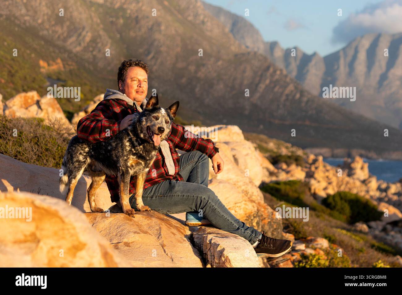 Ein Mann, der draußen sitzt und seinen australischen Rinderhund (Blue heeler) hält, wandert und erkundet zusammen mit Blick auf die Berge, und blickt in die Ferne Stockfoto