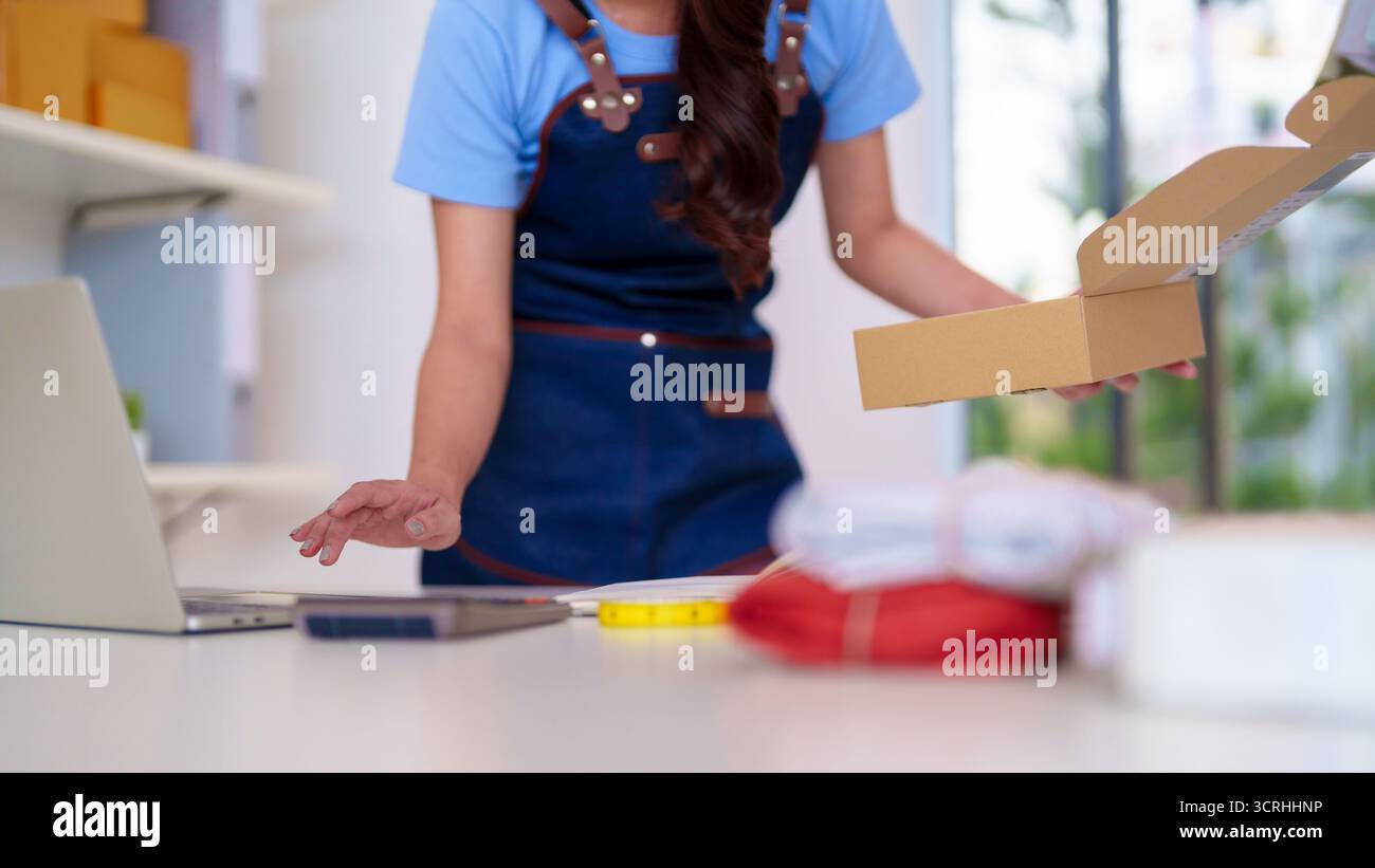 Eine Erwachsene asiatische Unternehmerin verpackt sorgfältig Bekleidungsbestellungen für ihr kleines Unternehmen, die Artikel an Kunden versenden, die die Logistik von ihrem Zuhause aus verwalten Stockfoto