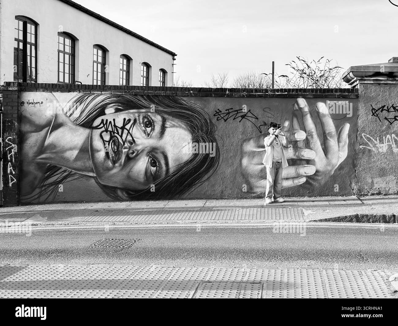 Street Art Wandgemälde mit einem detaillierten Porträt, teilweise mit Graffiti bedeckt, während eine Person davor steht und die Szene mit einer Kamera festnimmt Stockfoto