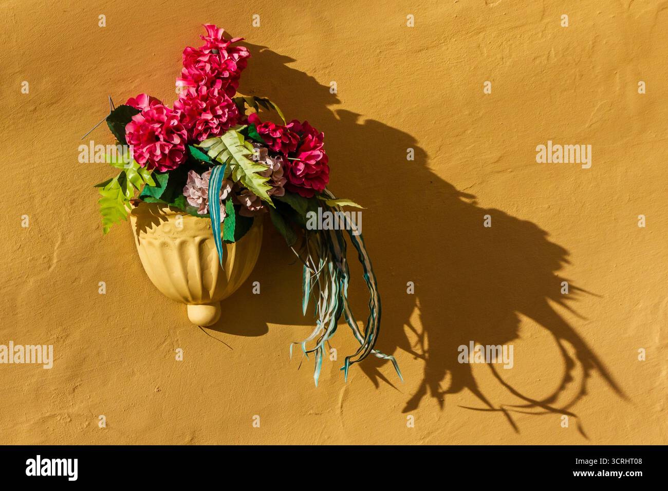 Rosafarbenes Blumenarrangement in keramischem Wandübertopf gegen sonnige gelbe Wand mit starkem Schatten Stockfoto