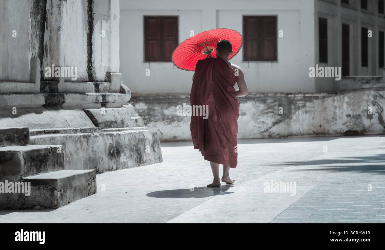 Buddhistischer Mönch mit rotem Regenschirm neben der antiken Tempelmauer in Myanmar, friedlicher spiritueller Moment in sanftem Licht Stockfoto