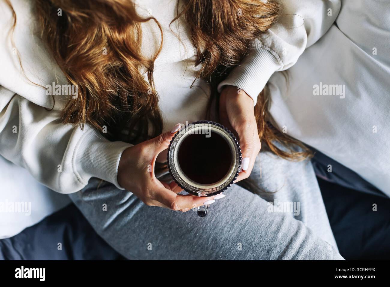 Frau in einem gemütlichen Pullover, der eine Tasse Kaffee hält. Entspannte Morgenroutine zu Hause. Warmes Getränk und friedlicher Lifestyle-Moment. Komfort und ruhige Atmosphäre im Innenbereich. Stockfoto