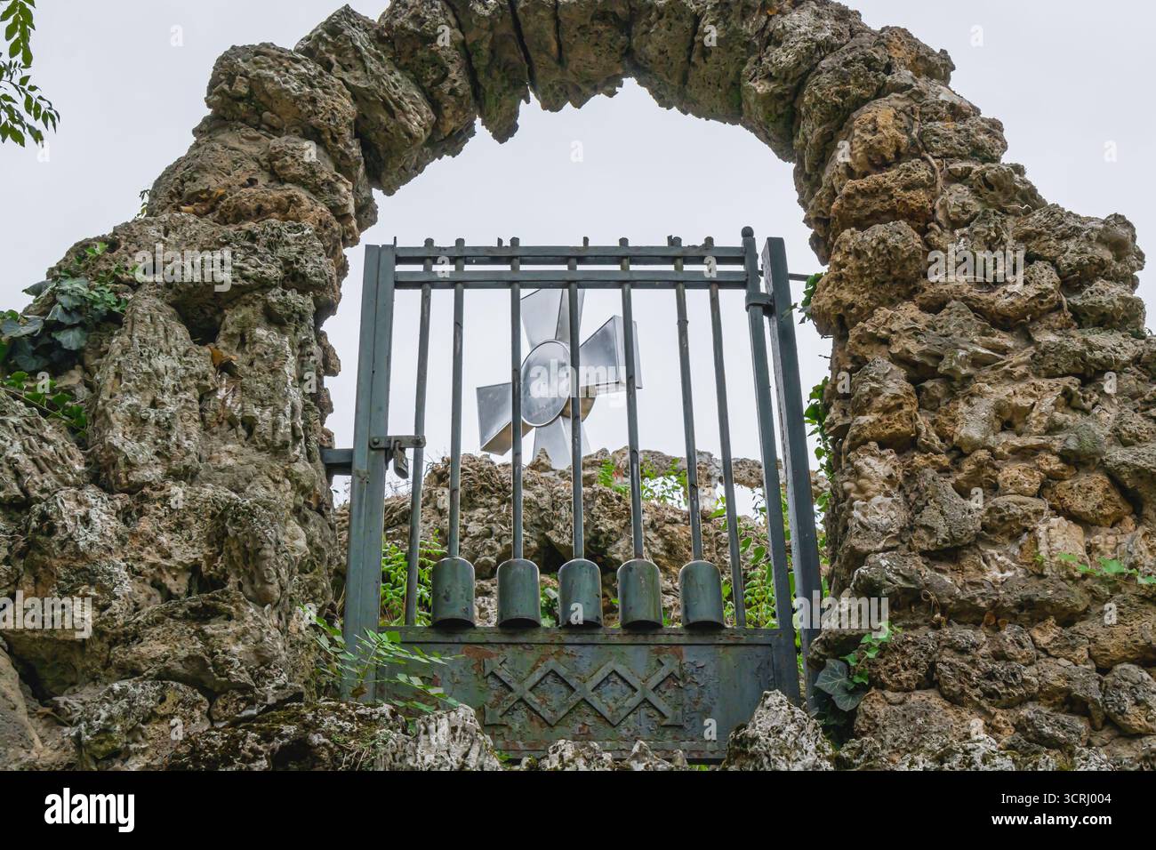 Ein alter Steinbogen mit einem verwitterten Metalltor und Kreuz-Symbol, eingerahmt von zerklüfteten Felsen und Grün, erinnert an Geschichte, Glauben und zeitlose Widerstandsfähigkeit. Stockfoto