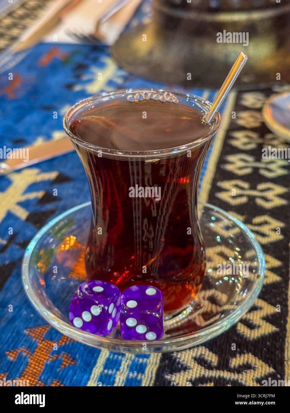 Türkisches Teeglas mit lila Würfeln auf farbenfrohen gemusterten Tisch für eine warme, verspielte Atmosphäre Stockfoto