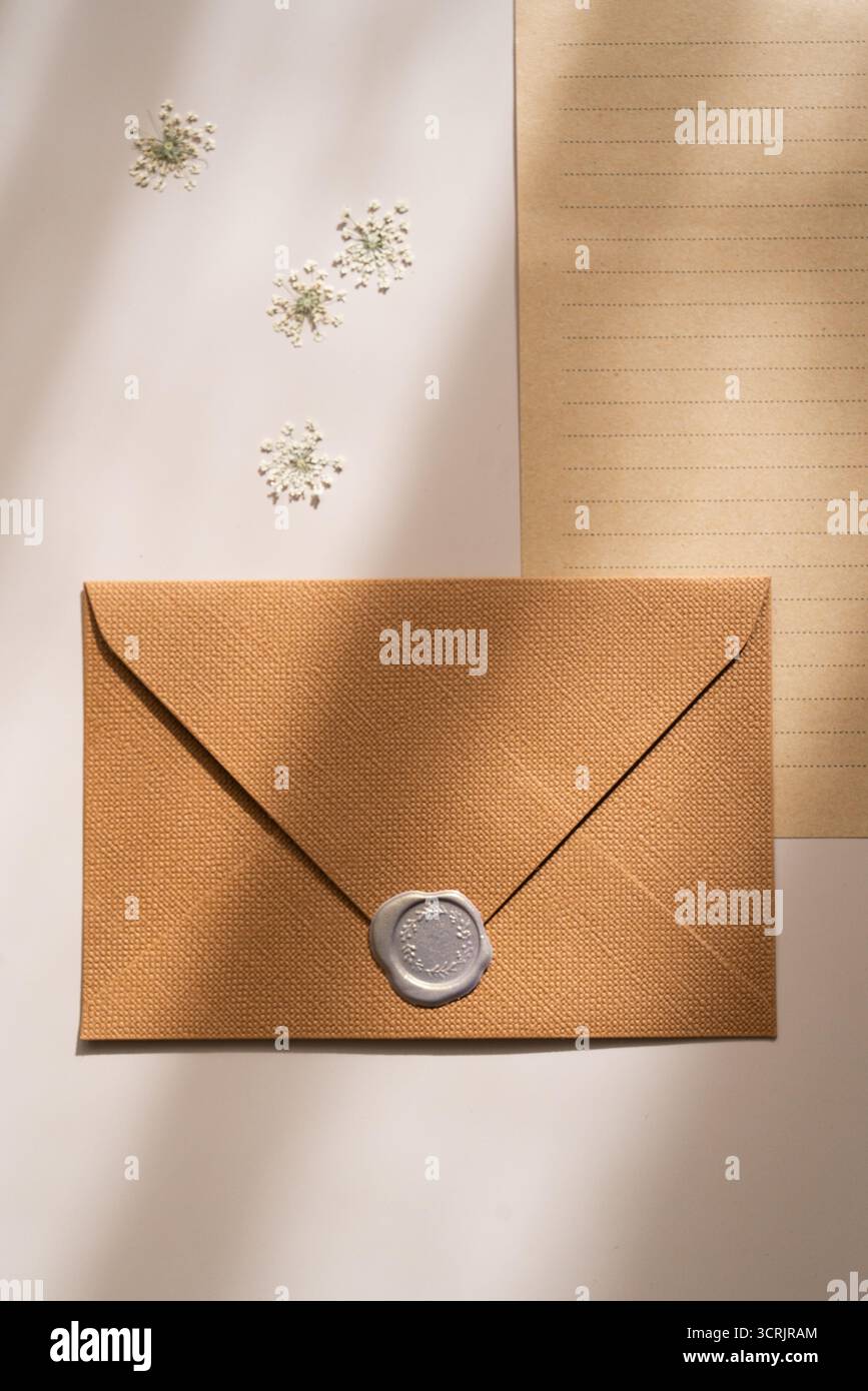 Kraftumschlag mit grauem Wachssiegel und liniertem Papier, zarte Blüten, weiche Schatten, flache Lagen Stockfoto