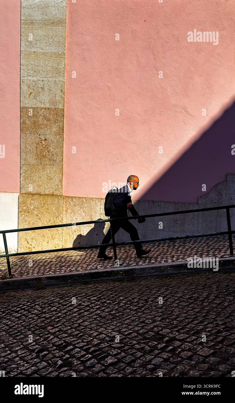 Mann schreitet zügig über sonnenbeschienene Kopfsteinpflaster, Schatten einer rosa Wand, städtische Straßenszene, Lissabon Stockfoto