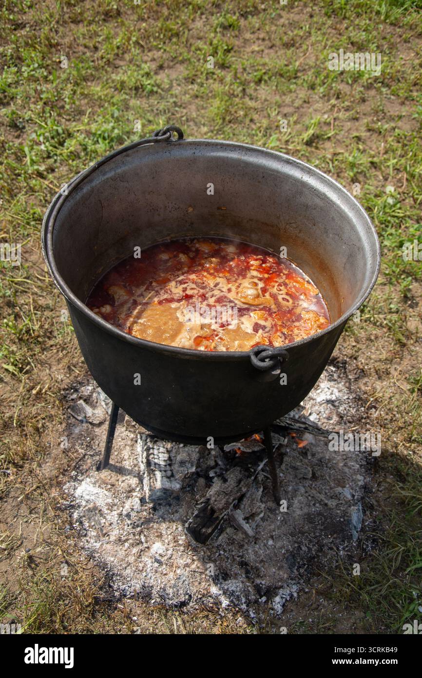 Ein großer Topf, wie ein Kessel, auf einem Stativ über einem offenen Feuer, in dem eine Schüssel zubereitet wird. Das scheint ein Gulasch (Gulyás) zu sein, eine deftige Suppe Stockfoto