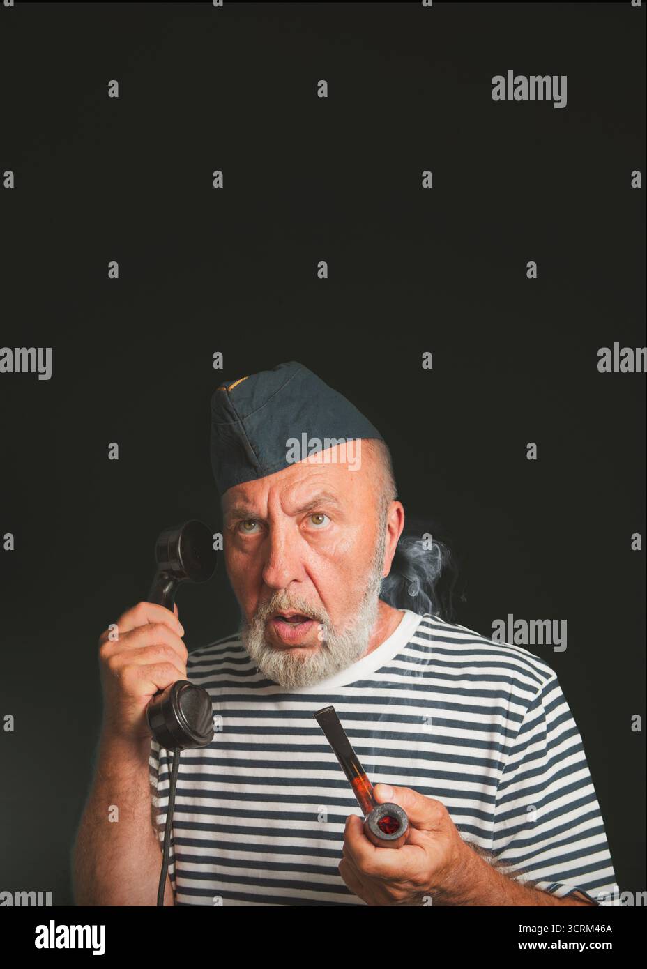 Porträt eines authentischen Seemanns aus der Vergangenheit spricht am Telefon. Auf schwarzem Hintergrund. Ein alter Seemann mit einer rauchenden Pfeife bringt Überraschung auf dem Ausdruck Stockfoto