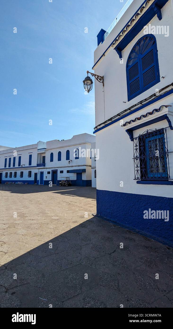Lebendige Straßenszene in Asilah, Marokko, mit weiß getünchten Wänden, dunkelblauen Fensterläden und traditionellen Laternen unter hellem mediterranem Sonnenlicht - Smartphone-aufgenommenes Stockfoto
