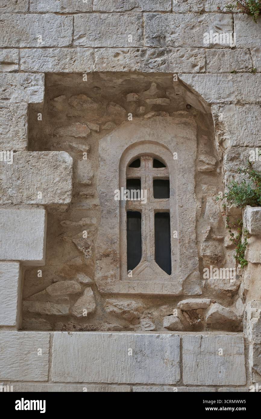 Nahaufnahme der mittelalterlichen Steinmauer mit Bogenfenster und geschnitztem Kreuz, Detail der historischen christlichen Architektur in Europa Stockfoto