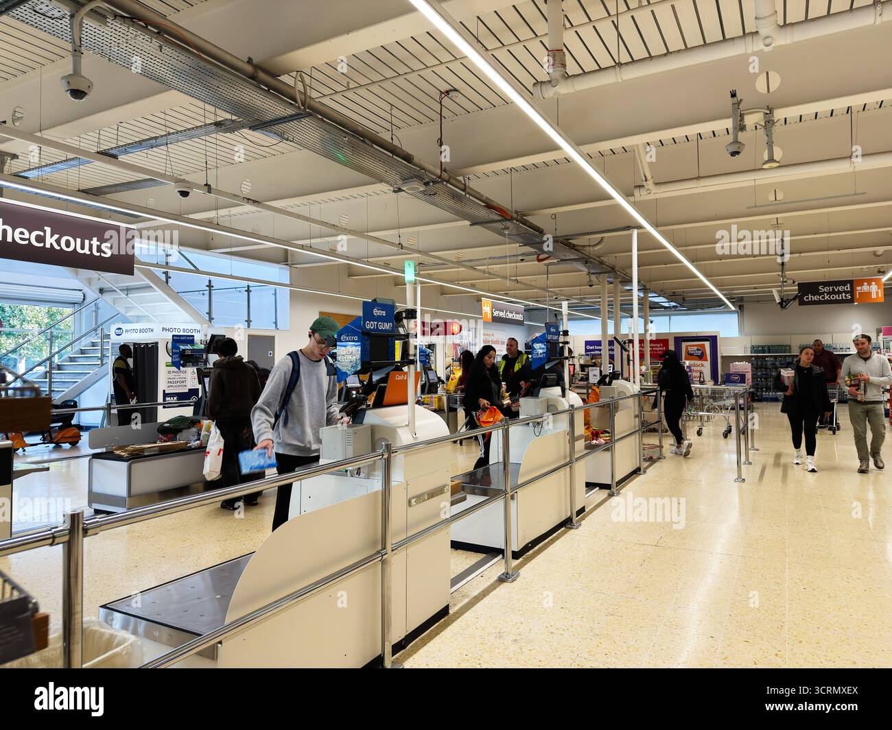 Salford, Großbritannien - 25. September 2025: Käufer nutzen Selbstkasse, während die Mitarbeiter an den Kassen in einem modernen, hellen Sainsbury's Geschäft helfen. Stockfoto