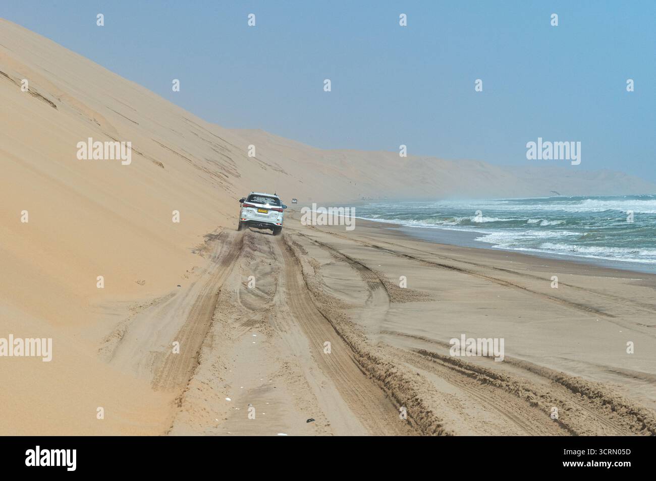 Weißes Geländefahrzeug, das entlang der Atlantikküste in Sandwich Harbour, Namibia fährt und Reifenspuren auf nassem Sand mit nebeligem Strand und Dünen hinterlässt Stockfoto
